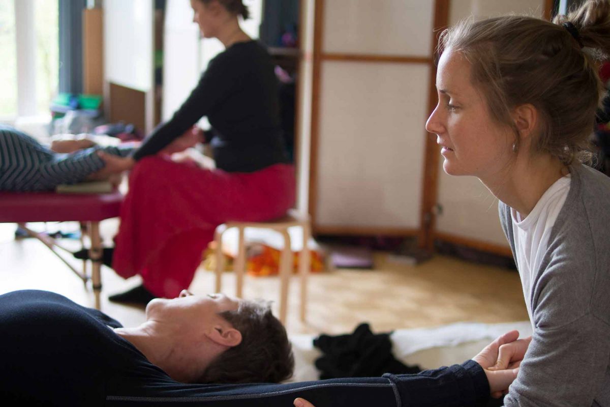 Im Vordergrund liegt eine Person, während eine zweite Person ihren Arm hält und dehnt. Im Hintergrund sitzt eine weitere Person auf einem Hocker und ist ebenfalls in eine körperliche Interaktion mit einer teilweise sichtbaren Person eingebunden.
